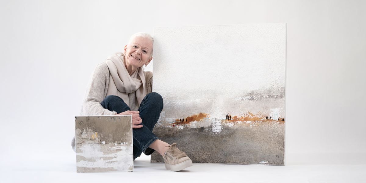 Künstlerin Sigrid Koller sitzt im Schneidersitz auf dem Boden, vor sich das Bild "Aschegold" und lehnt lächelnd ihre Stirn an die Leinwand des Bildes "Toskana".